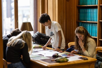 three young students sat studying at a table with a male student stood over his papers